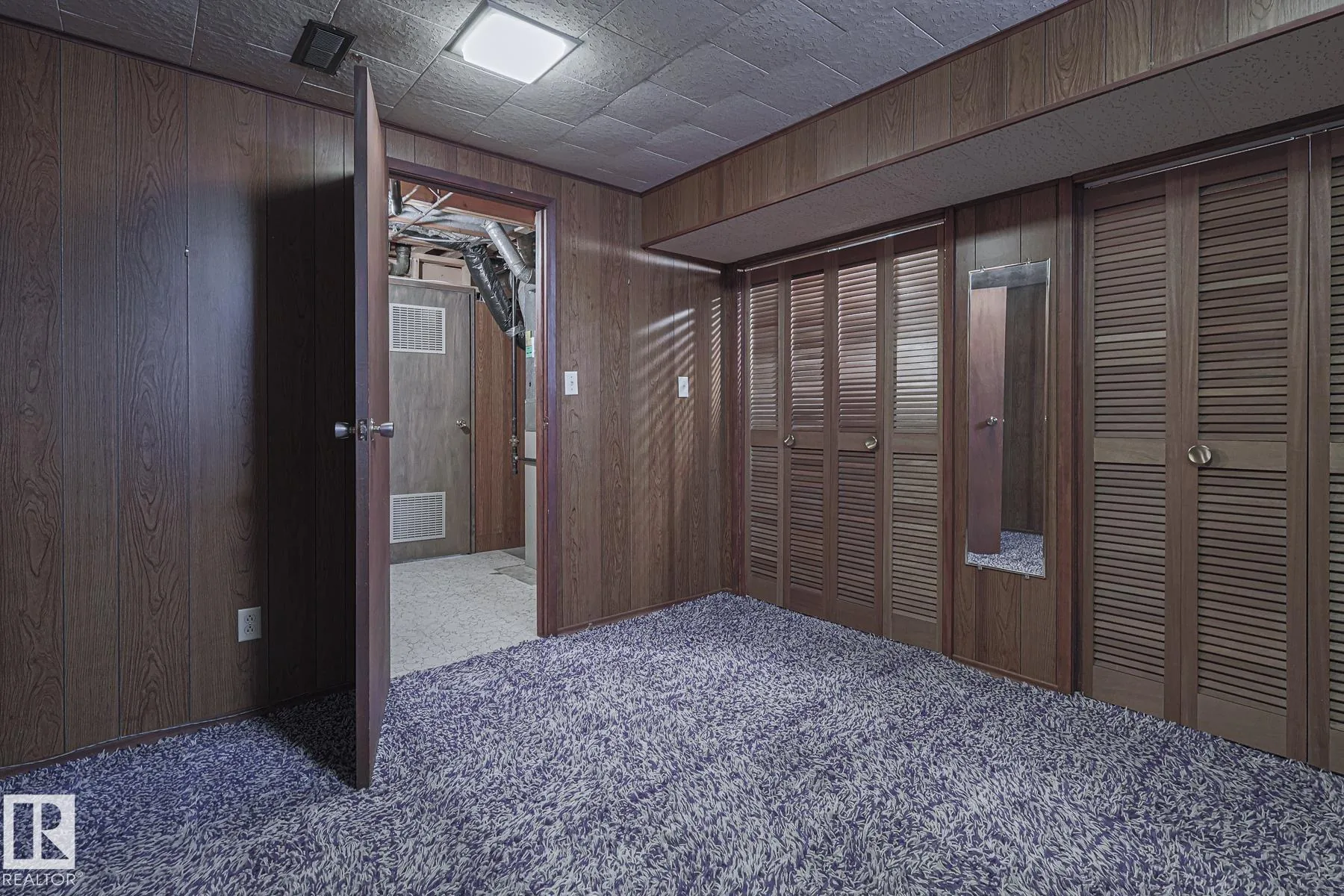 Before renovation, another angle of the basement bedroom showing louvered closet doors, a narrow wall mirror, and a plywood door open toward the mechanical/utility room.