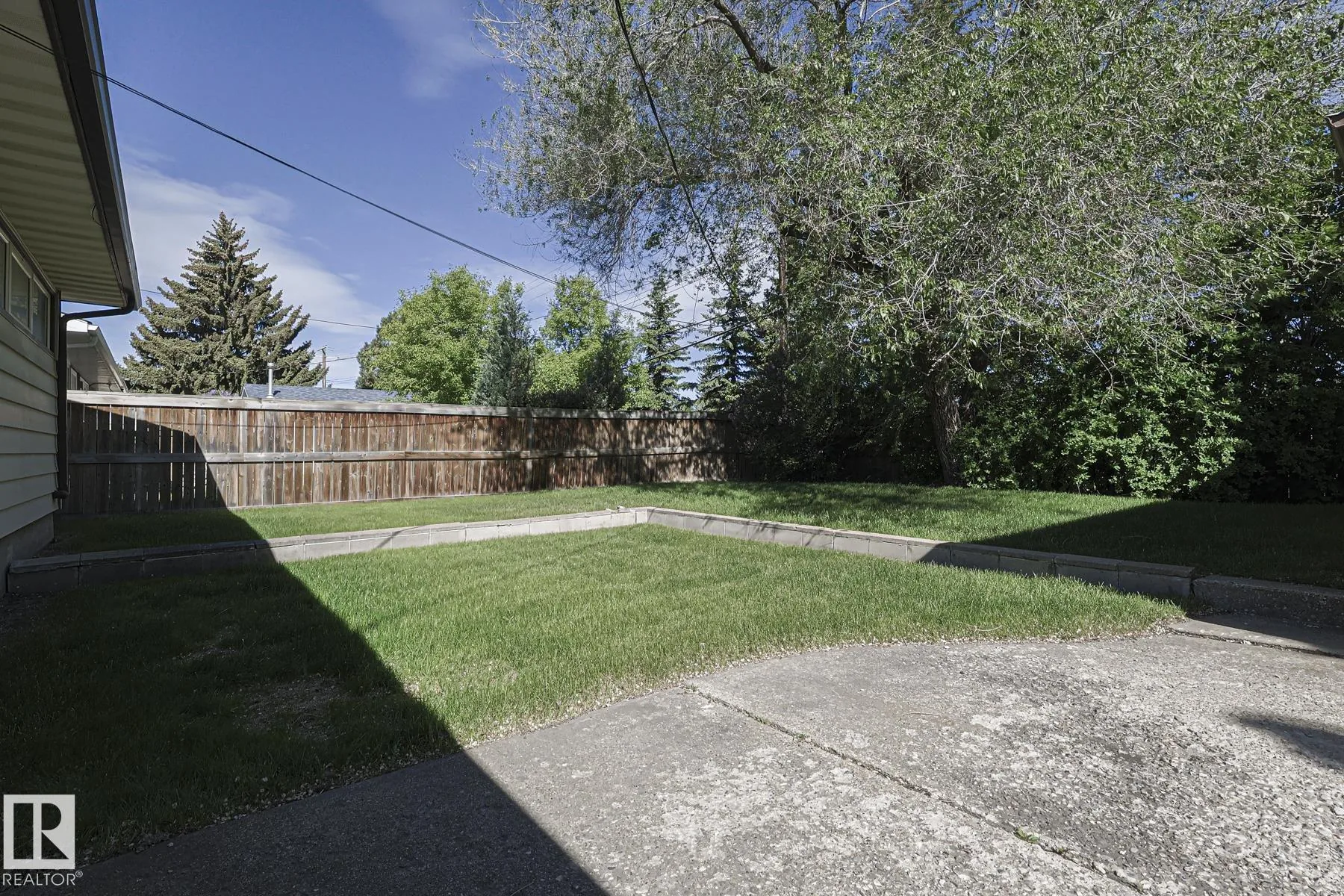 Before renovation, the backyard corner with a low concrete-block planter edging the lawn, wooden fence at the rear, and mature trees overhanging.