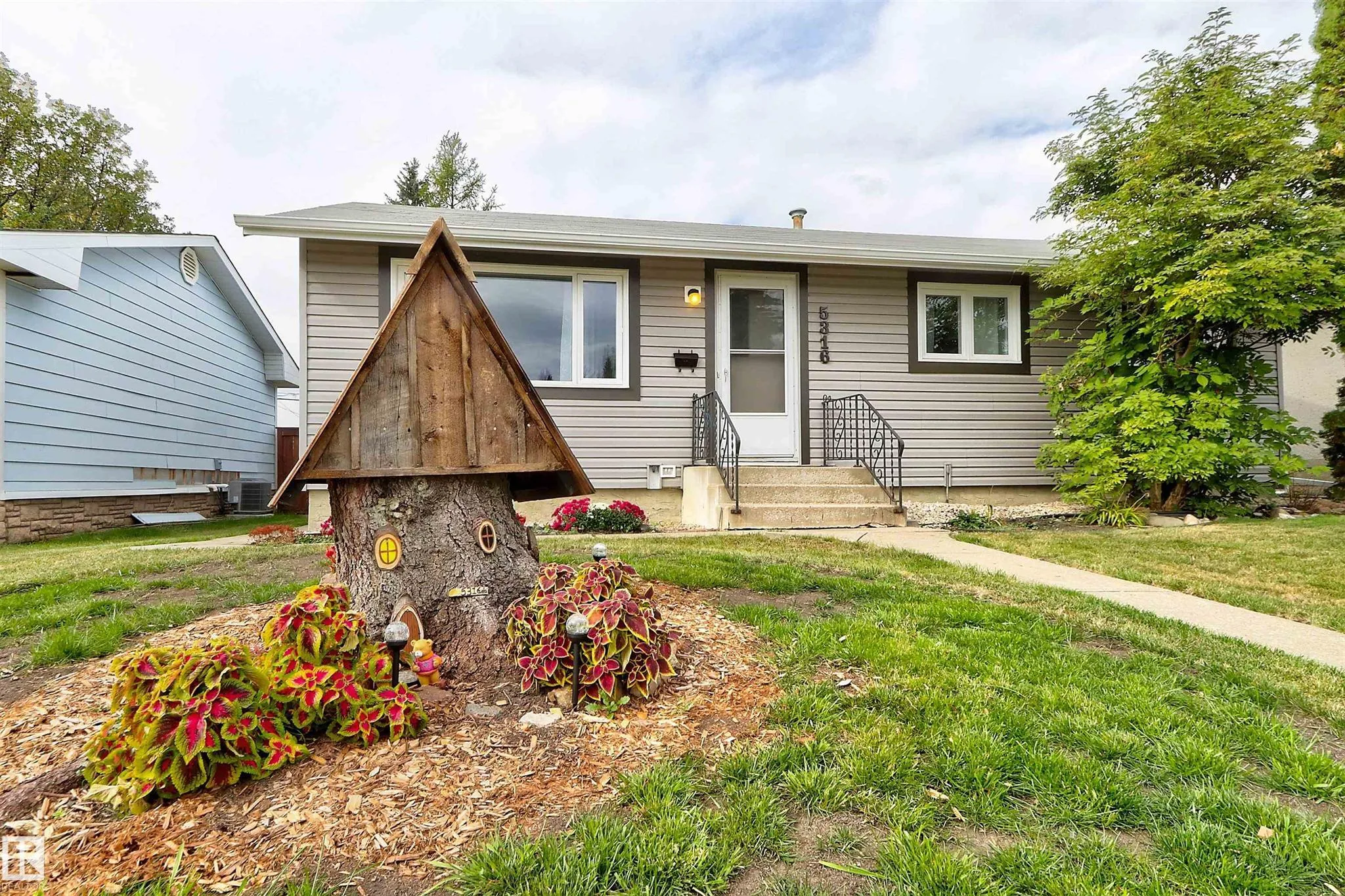Before renovation, closer front-elevation shot of the bungalow with the wrought-iron railed porch, a tree-stump fairy-house decoration by the walk, and the 5316 house number.