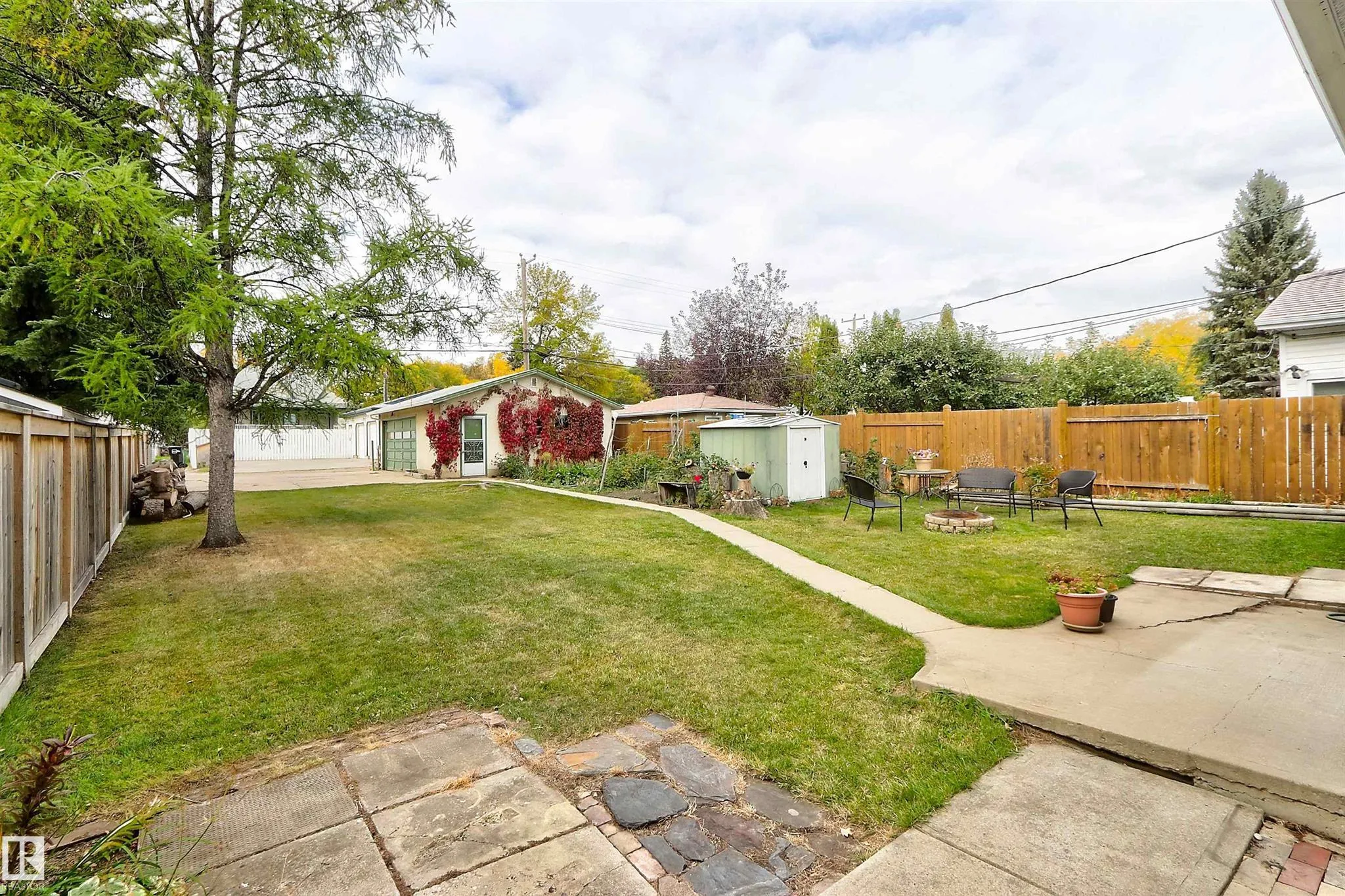 Before renovation, wide backyard view toward the detached stucco garage covered in red Virginia creeper, with a large evergreen and iron-frame patio seating.