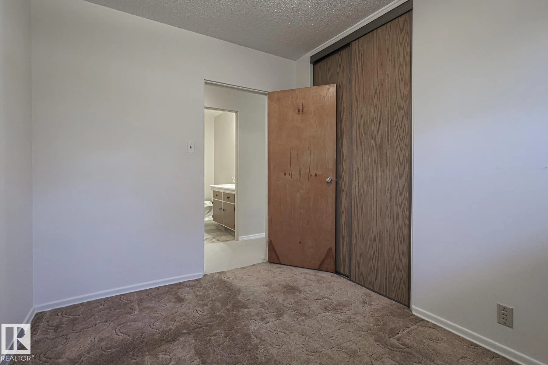 Before renovation, a small bedroom with brown patterned shag-style carpet, a plywood swing door, and a woodgrain sliding closet on the right; bathroom vanity visible through the open doorway.