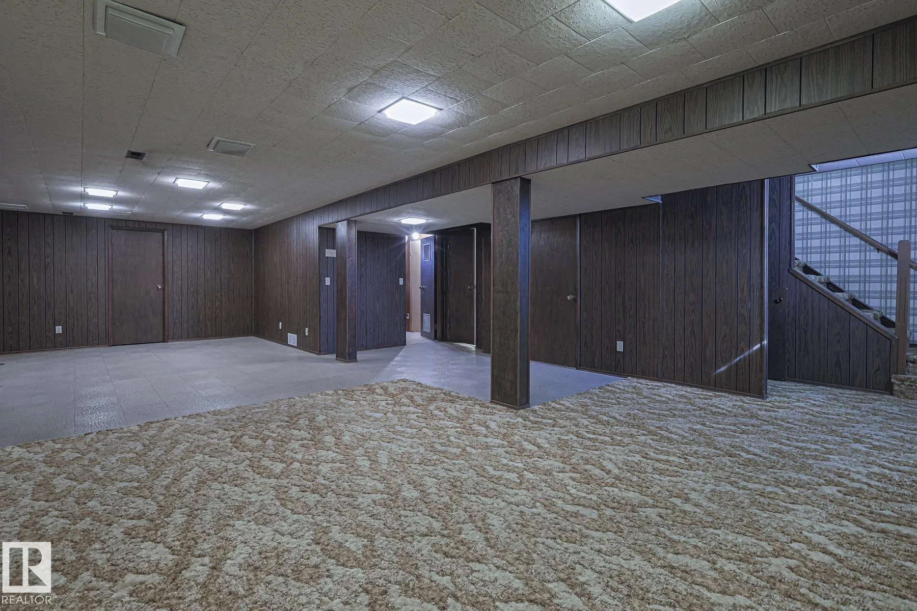 Before renovation, the basement recreation area with dark faux-wood paneled walls, a drop-tile ceiling, mixed vinyl-tile and cream-tone shag carpet flooring, and stairs at right.