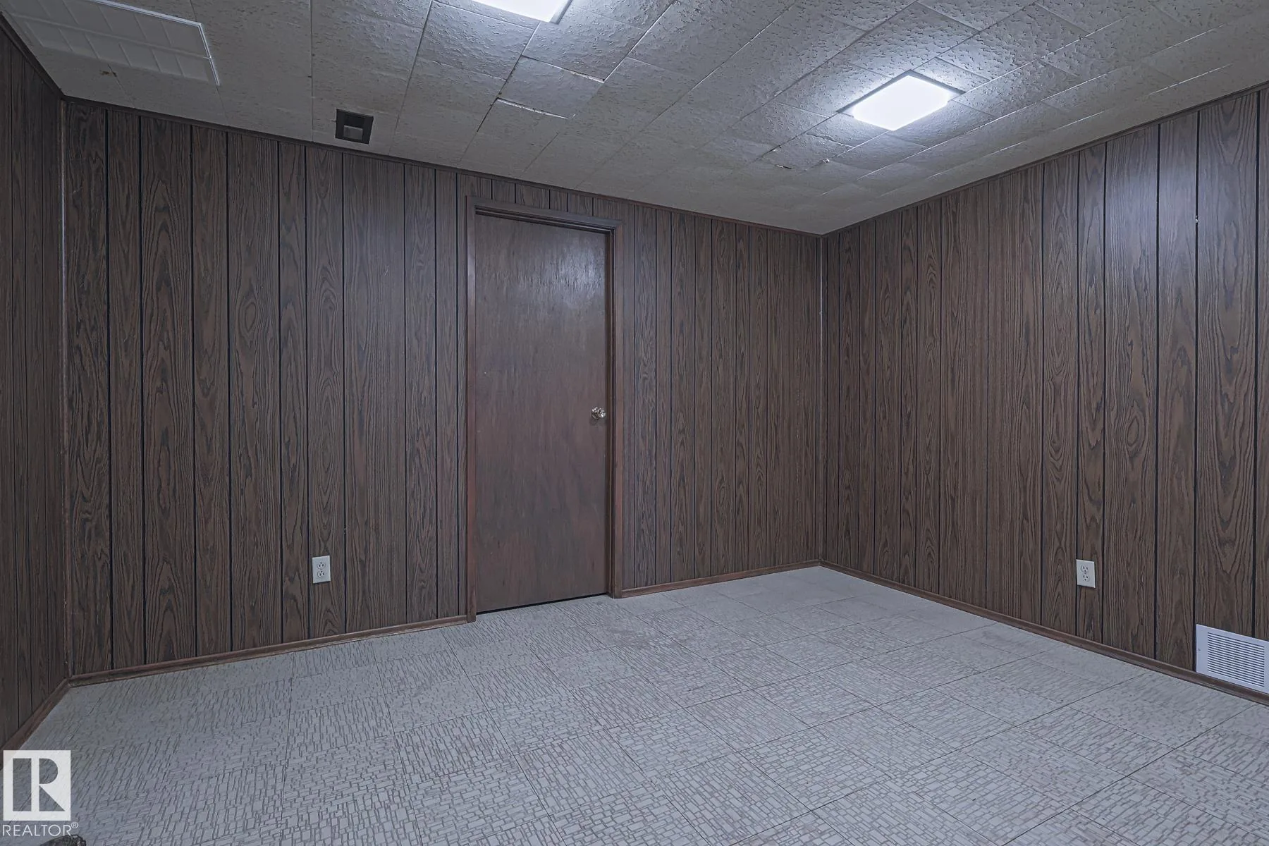Before renovation, a basement bedroom with dark faux-wood paneled walls, purple-and-cream shag carpet, and a small casement window with sheer curtain near the ceiling.