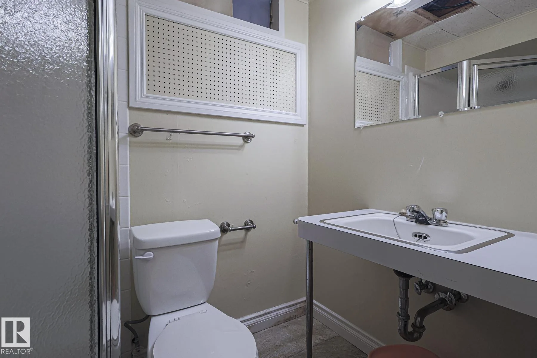 Before renovation, the basement bathroom with a fiberglass shower stall with textured obscure-glass doors, a wall-mounted sink with exposed plumbing, a toilet, and a pegboard vent panel above.