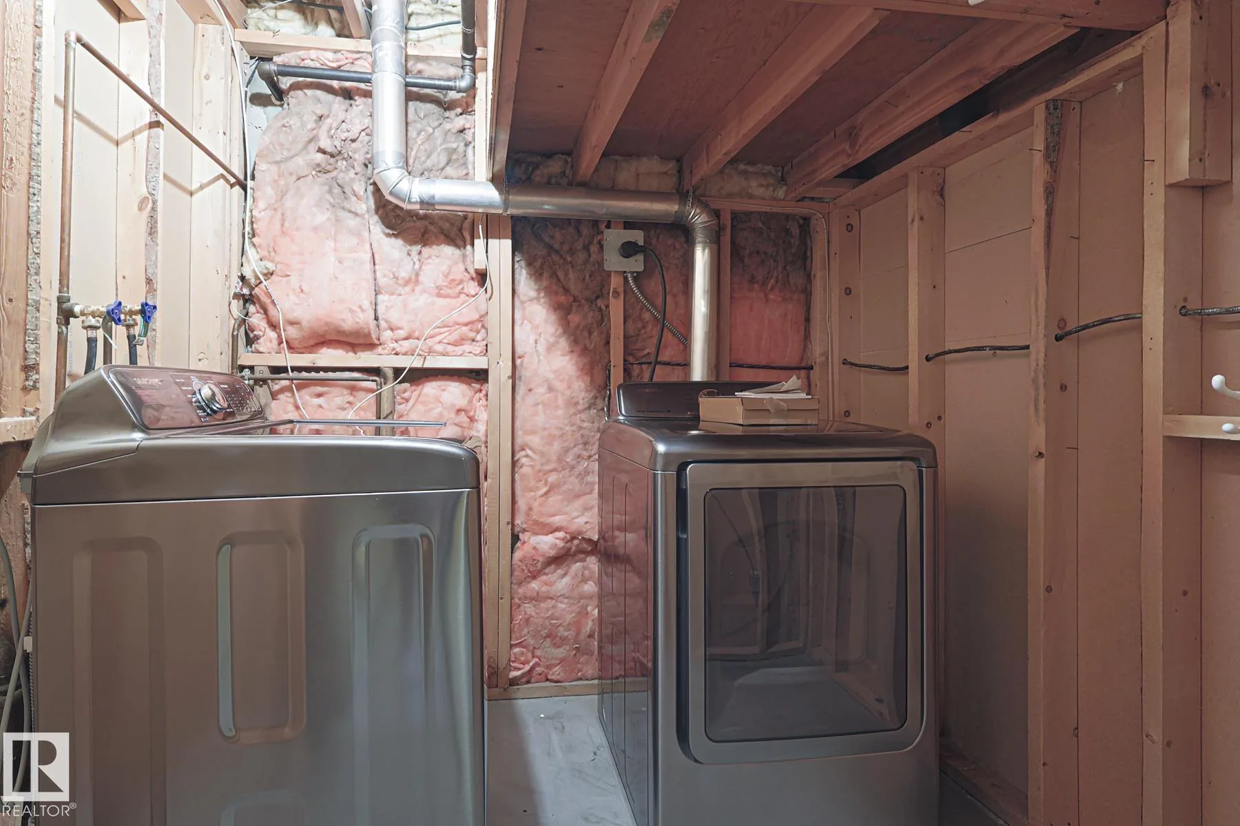 Before renovation, the laundry area in an unfinished framed section of the basement with a champagne-color top-load washer and matching front-load dryer against exposed pink insulation and pine studs.